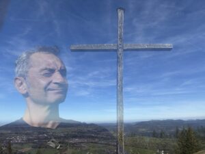 Ein durchscheinendes Männergesicht erscheint neben einem großen Holzkreuz auf einer Bergkuppe